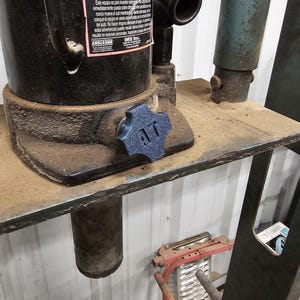 Op de afbeelding: Een close-up van een zwarte hydraulische krik met een blauwe knop met de letters "AT". De krik heeft waarschuwingslabels in het Engels en Spaans. De krik staat op een metalen oppervlak.