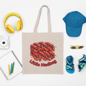 May include: A beige tote bag with a graphic of red crawfish and the text "Little Mudbug." The image also includes a blue baseball cap, yellow headphones, a white camera, sunglasses, sandals, and a notepad with crayons.