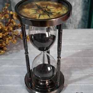 May include: An antique-style hourglass with a compass design on top. The hourglass has a dark bronze finish and contains black sand. The base and top are circular, and the glass is clear. The hourglass is a decorative item.