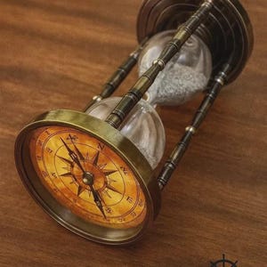 Op de afbeelding: Een zandloper in antieke stijl met een kompasontwerp. De zandloper heeft een messingkleurig frame en een heldere glazen bol gevuld met zand. De kompasroos is gedrukt op een gele achtergrond. De tekst "ZIYA NAUTICAL" is zichtbaar.