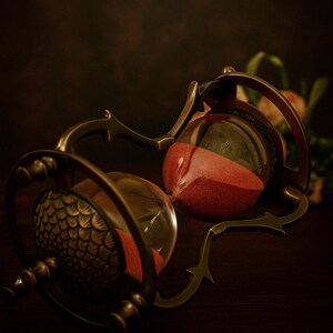 May include: An antique-style hourglass with a bronze-colored frame and red sand. The glass bulbs are connected by a decorative metal structure. The text "Ziya Nautical" is displayed at the top.