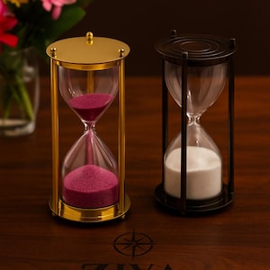 May include: Two hourglasses, one gold-toned with pink sand and the other black with white sand, sit on a wooden surface. The hourglasses are part of the ZIYA NAUTICAL collection. A vase of flowers is in the background.