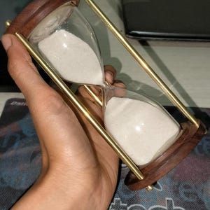 May include: An hourglass featuring two clear glass bulbs filled with white sand, connected by four slender brass-toned metal rods and secured at both ends by circular dark brown wooden caps with a smooth, polished finish.