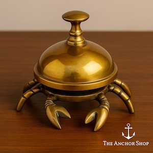 May include: A polished brass desk bell shaped like a crab with a rounded dome top and a central handle. The crab features six articulated legs and two front claws. The text "THE ANCHOR SHOP" appears in the bottom right corner with an anchor icon.