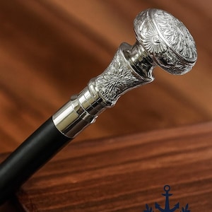 May include: Silver-coloured walking cane with a black shaft. The handle features intricate floral and scrollwork designs. The cane's top is a rounded, decorative cap. The ZIYA NAUTICAL logo is visible in the background.