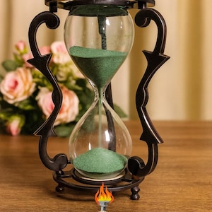 May include: An ornate hourglass with green sand, held within a black metal frame. The glass bulb is supported by decorative, curved side supports. The base has the text "ZIYA NAUTICAL".