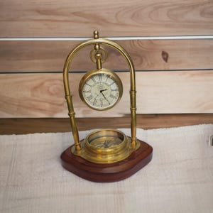 May include: A brass desk clock with a compass, set on a wooden base. The clock face is cream-coloured with black Roman numerals and hands. The compass is visible below the clock. The clock is suspended within a brass arch.
