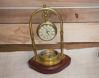 May include: A brass desk clock with a compass, set on a wooden base. The clock face is cream-coloured with black Roman numerals and hands. The compass is visible below the clock. The clock is suspended within a brass arch.
