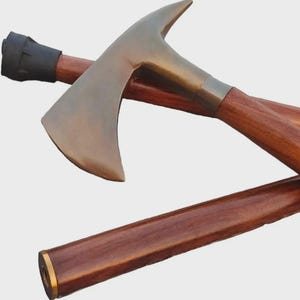 Puede incluir: Hacha con cabeza de color bronce y mango de madera. La cabeza del hacha tiene una hoja curva y una parte superior puntiaguda. El mango es de color marrón oscuro con un agarre de goma negra.