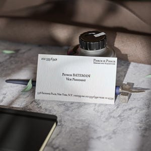 以下が含まれることがあります： 「Patrick Bateman」の名前と「Vice President」の肩書きが書かれた白い名刺。 ペンと金属製のオブジェクトで支えられています。 インクボトルとノートも表示されています。 テキストは黒です。