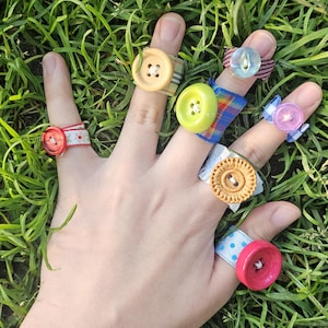 Puede incluir: Una mano que lleva siete anillos de botón de varios colores y diseños. Los anillos presentan botones en tonos de rojo, amarillo, verde y azul, sujetos a bandas de tela con patrones como lunares y rayas. La mano está sobre un fondo de hierba verde.