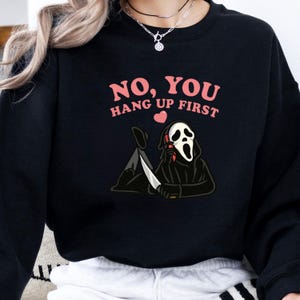 May include: Black sweatshirt with the text "NO, YOU HANG UP FIRST" in pink. The graphic features a cartoon figure in a black hooded robe and a white mask, holding a knife and a phone. A pink heart is above the figure.