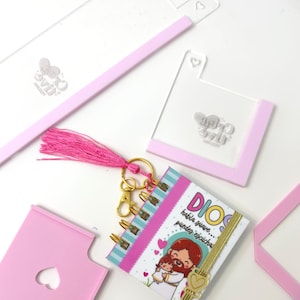 May include: A selection of pink and clear acrylic accessories, including a keychain with a small notebook. The notebook features a colourful illustration and the text "Dios habla suave... puedes escucha". A pink tassel and gold-coloured hardware are also present.
