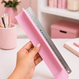 May include: A light pink plastic ruler with a heart-shaped end and a raised edge, featuring a metal insert with black measurement markings ranging from 1 to 30 centimeters, held to display the scale against a plain white background.