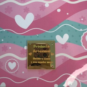 May include: A decorative background with a repeating pattern of pink and teal waves, white hearts, and floral accents. A gold-colored metal tag is affixed to the surface, with the words "Producto Artesanal" and other text.