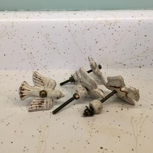 May include: A collection of white ceramic bird-shaped cabinet knobs with brown accents. Each knob has a metal screw for installation. The knobs are arranged on a speckled white surface, suggesting a home decor theme.