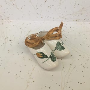 May include: A pair of white ceramic clogs with a floral design featuring yellow and green accents. The clogs are adorned with a brown ribbon bow. These decorative clogs are a charming addition to any home decor collection.