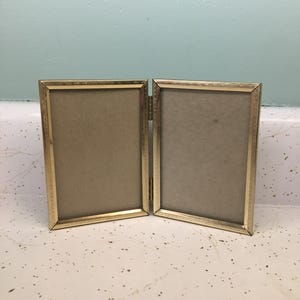 May include: A hinged, gold-colored metal picture frame with two rectangular openings. The frame is open, revealing a tan backing in each section. The frame is sitting on a speckled white surface.