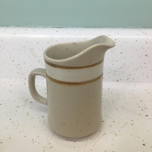 May include: A beige ceramic creamer with a curved spout and a small handle. The creamer has a light brown band around the middle and a speckled finish. The creamer is likely part of a vintage dinnerware set.