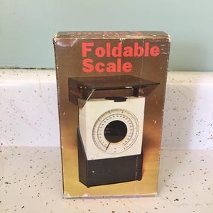 May include: A vintage foldable scale in its original box. The box is brown with red text that reads "Foldable Scale." The scale itself is white with a black base and a circular dial. The dial has measurements in ounces and pounds.