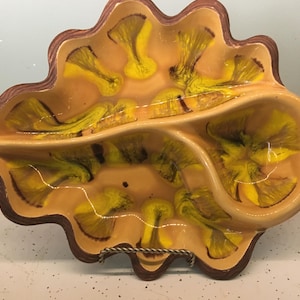 May include: A vintage, leaf-shaped serving dish with three compartments. The dish features a brown rim and a tan interior with yellow and green abstract designs. It is supported by a metal stand, perfect for serving snacks or appetizers.