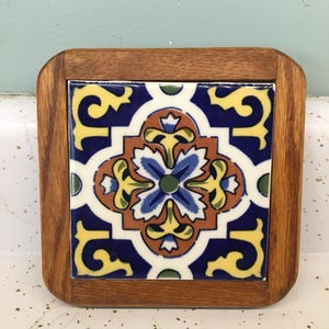 May include: Square wooden coaster with a colorful ceramic tile insert. The tile features a floral design with blue, yellow, orange, and green accents on a white background. The wooden frame has a natural finish.