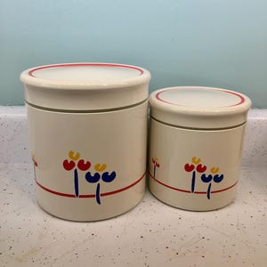 May include: Two off-white ceramic canisters with lids. Each canister has a red, yellow, and blue floral design and a red stripe. The lids have a red trim. The canisters are different sizes.