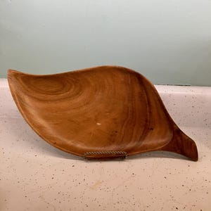 May include: A wooden leaf-shaped serving dish with a natural wood grain finish. The dish has a curved design and a handle-like extension. The dish is approximately 15 inches long.