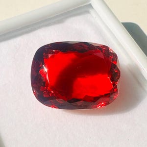 May include: A faceted, cushion-cut gemstone with a deep red hue. The gem is set in a white display case, showcasing its brilliance. The stone's surface reflects light, highlighting its intricate cuts and vibrant color.