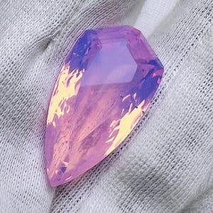 May include: A faceted, elongated gemstone with a gradient of colors. The gem transitions from a pale yellow at the base to a vibrant pink and purple at the top. The stone is set against a white fabric background.