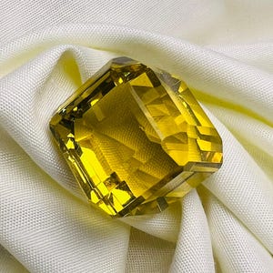 Puede incluir: Una gema facetada de color amarillo dorado con forma rectangular. La gema se encuentra sobre una tela blanca, creando un contraste que resalta su brillo. El corte de la piedra revela múltiples facetas, realzando su brillo y atractivo visual.
