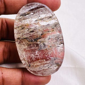May include: An oval-shaped, translucent gemstone with internal inclusions of brown, black, and reddish-pink hues. The stone has a polished surface, and the inclusions create a layered, textured appearance. The gemstone is held in a hand against a white background.
