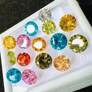May include: A collection of round, faceted gemstones in various colors, including yellow, blue, pink, orange, and green, displayed in a white tray. The gemstones are arranged closely together, showcasing their brilliance and clarity.