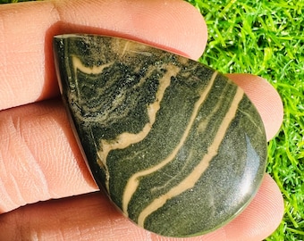 May include: A polished, teardrop-shaped gemstone with a dark green base and swirling, light tan bands. The stone is held in a hand, showcasing its smooth, reflective surface. The natural patterns create a unique, organic design.
