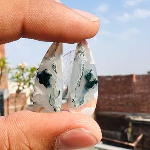 Puede incluir: Dos piedras preciosas de ágata musgosa pulidas, cada una con forma de lágrima. Las piedras muestran una base blanca translúcida con intrincados patrones de verde, negro y toques de naranja. Las piedras preciosas se sostienen entre los dedos contra un cielo azul.