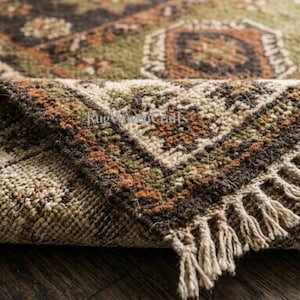 May include: A woven area rug featuring a folded corner that reveals a textured surface with a geometric pattern in shades of olive green, dark brown, and burnt orange. The rug has a cream-colored fringe along the edge and the text RugWoodCrafts.