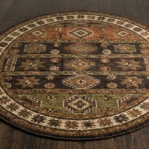 May include: A round area rug featuring a multi-colored geometric pattern with horizontal bands in shades of brown, dark charcoal, and muted green. The border displays a repeating star and diamond motif in cream, tan, and dark brown tones.
