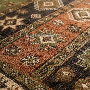 May include: A woven rug featuring a repeating geometric pattern in shades of olive green, burnt orange, black, and cream. The surface has a textured, looped pile construction with intricate star and diamond motifs arranged in horizontal bands.