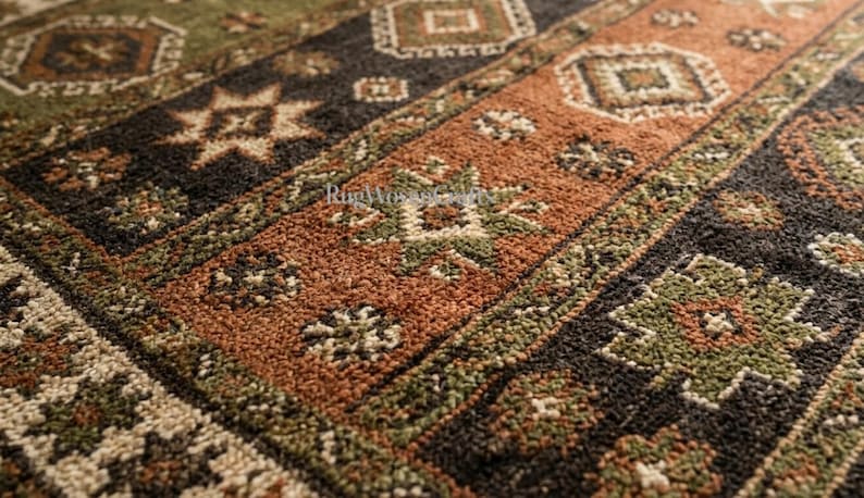 May include: A woven rug featuring a repeating geometric pattern in shades of olive green, burnt orange, black, and cream. The surface has a textured, looped pile construction with intricate star and diamond motifs arranged in horizontal bands.