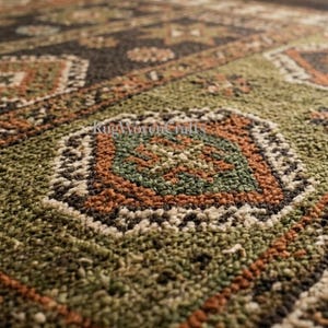 May include: A textured area rug featuring a repeating geometric pattern in shades of olive green, dark brown, cream, and burnt orange. The surface has a thick, looped pile construction with distinct diamond-shaped motifs and linear borders.