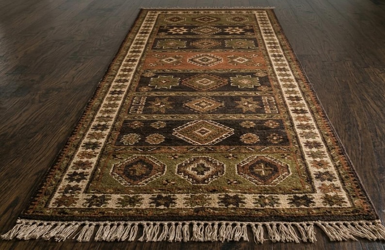May include: A rectangular area rug featuring a series of horizontal bands in shades of dark brown, olive green, and terracotta. The rug is decorated with repeating geometric diamond and star patterns, finished with a cream-colored border and fringe.