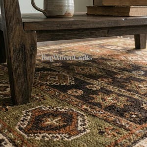 May include: A rectangular area rug featuring a repeating geometric pattern in shades of olive green, dark brown, and burnt orange. The rug is positioned beneath a dark brown wooden coffee table with a ceramic mug and two stacked books on top.