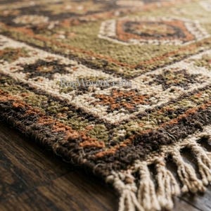 May include: A rectangular area rug featuring a woven pattern in shades of dark brown, olive green, and cream. The rug has a decorative border with geometric star-like motifs and a fringe edge made of thick, light-colored twisted yarn tassels.