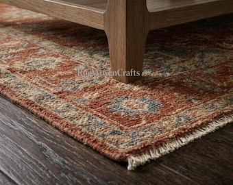 May include: A rectangular area rug featuring a detailed pattern in shades of terracotta, muted blue, and cream. The rug has a short fringe along the edge and is positioned beneath the wooden legs of a piece of furniture on a dark wood floor.