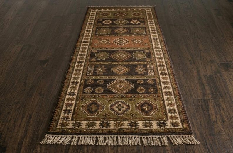 May include: A rectangular runner rug featuring a series of horizontal panels with geometric diamond and star motifs in shades of olive green, dark brown, and terracotta. The rug is framed by a cream border and finished with light beige fringe.