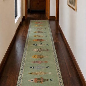 May include: A long, rectangular runner rug in a soft sage green colour, featuring a repeating pattern of stylised fish in various colours and textures arranged in a single vertical row, finished with a decorative border and white fringe at the ends.