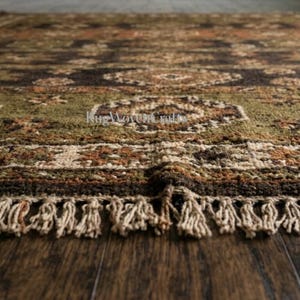 May include: A rectangular area rug featuring a complex geometric pattern in shades of olive green, dark brown, rust orange, and cream. The rug has a thick, textured pile and is finished with a row of cream-colored fringe along the bottom edge.
