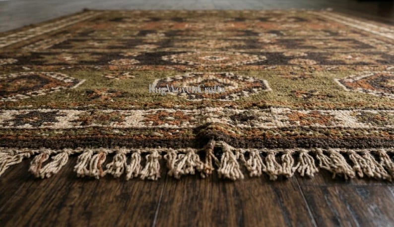 May include: A rectangular area rug featuring a complex geometric pattern in shades of olive green, dark brown, rust orange, and cream. The rug has a thick, textured pile and is finished with a row of cream-colored fringe along the bottom edge.