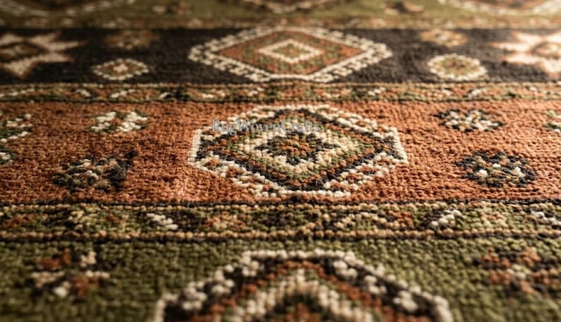 May include: A woven rug featuring a textured pile with a repeating geometric pattern in shades of olive green, burnt orange, and dark brown. The design includes diamond-shaped motifs and small floral-like accents across the surface.