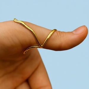 Può includere: Un anello color oro con un design unico a forma di V. L'anello si curva attorno al dito, con un'estremità che si estende sopra la nocca superiore. L'anello è su uno sfondo azzurro.